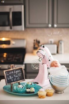 a baby in a kitchen next to a mixer and some cupcakes on a plate