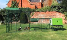 several chickens are in the grass near an outdoor chicken coop with a red brick building behind it