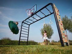 two children are playing in the grass near a basketball hoop and swing set that is attached to a wooden structure