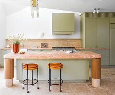 two stools sit at the center of a kitchen island with marble countertops and green cabinets