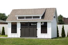 a white shed with black doors and windows on the roof is in front of a white picket fence