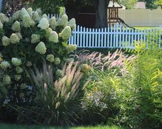 a white picket fence in front of a house with flowers and bushes around it,