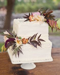 a white wedding cake with flowers and leaves on top sitting on a wooden table outside