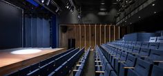 an empty auditorium with rows of blue seats and a wooden stage in the center is lit by spotlights