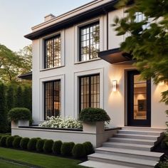 a white house with black windows and steps leading up to the front door is surrounded by greenery