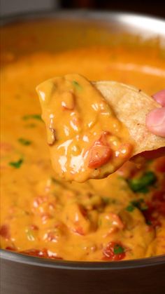 a hand holding a tortilla chip over a bowl of cheese soup with tomato sauce