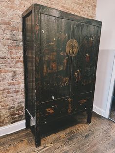 a black cabinet with paintings on it in front of a brick wall and wooden floor