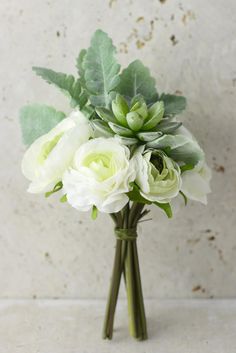 a bouquet of white flowers and green leaves in a vase on a counter top with a wall behind it