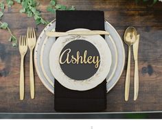 a place setting with black napkins and gold cutlery on the plate that says asley