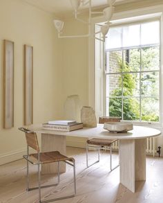 a room with a table, chairs and a book on the floor in front of a window