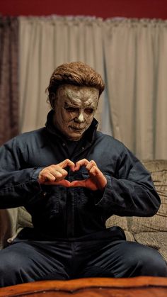 a man in a creepy mask sitting on a couch making a heart with his hands