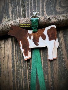 a brown and white cow shaped keychain hanging on a tree branch with green ribbon