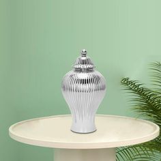 a silver vase sitting on top of a white table next to a potted plant