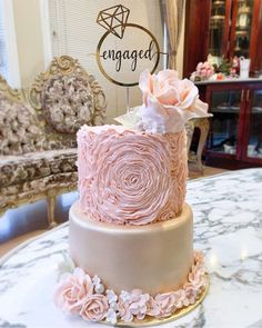 a wedding cake with pink flowers and engaged sign on the top tier, sitting on a marble table