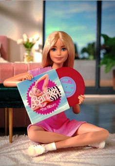 a barbie doll sitting on the floor holding a pink record in her hands and smiling