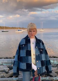 a woman standing on rocks near the water with her hand in her pocket, wearing a blue and white checkered coat