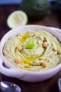 a white bowl filled with hummus next to two spoons and lime wedges