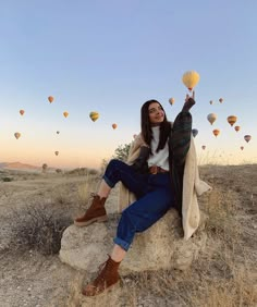 a woman sitting on top of a rock with hot air balloons flying in the sky