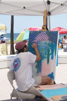 a man is painting on an easel in front of the beach with umbrellas