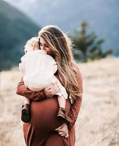 a woman holding a baby in her arms while standing on top of a grass covered field