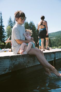 a woman holding a baby sitting on top of a wooden dock next to a body of water