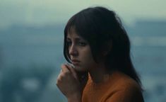 a woman in an orange sweater looking off into the distance with her hand on her chin