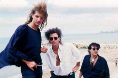 three men standing next to each other in front of the ocean and people on the beach