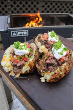 two loaded baked potatoes sitting on top of a wooden table next to an open fire pit