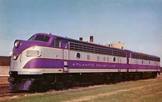 a purple and silver train traveling down tracks next to a grass covered field on a sunny day