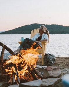 a woman sitting in a chair next to a campfire on the shore of a lake