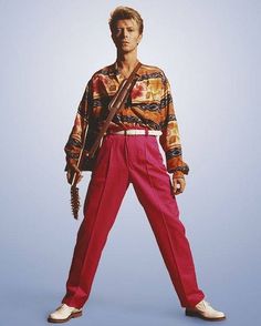 a man with a guitar standing in front of a blue background wearing red pants and a patterned shirt