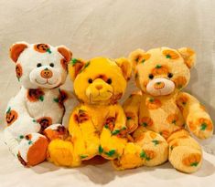 three stuffed teddy bears sitting next to each other on a white sheeted surface with orange and green decorations