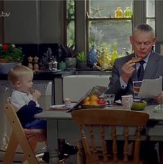 a man sitting at a table eating food with a little boy standing next to him