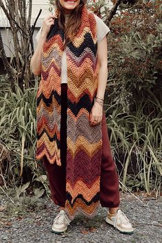 a woman standing in front of some bushes wearing a multicolored knitted scarf