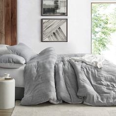 an unmade bed with grey linens in a white room next to two pictures on the wall