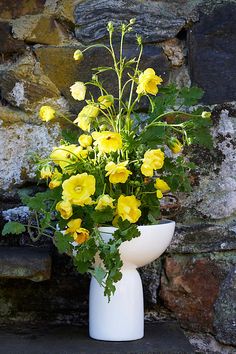 a white vase filled with yellow flowers next to a stone wall