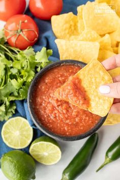 a person holding a tortilla chip with salsa and limes around the chips