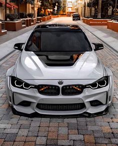 the front end of a white car parked on a cobblestone street with buildings in the background