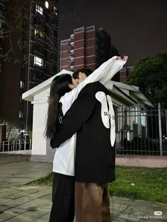 two people standing next to each other in front of tall buildings at night with their arms around each other