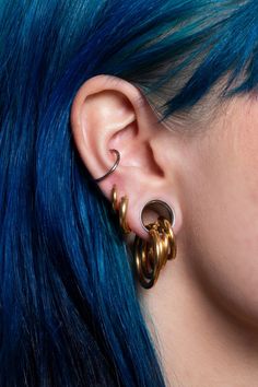 a woman with blue hair wearing gold ear cuffs and piercings on her ears is looking at the camera