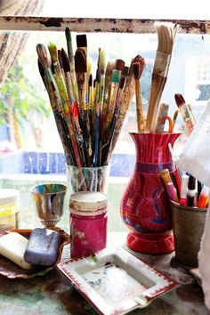 a vase filled with lots of paint and brushes on top of a table next to a window