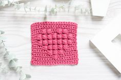 a pink crocheted square sitting on top of a white table next to flowers