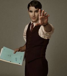 a man in a vest and tie holding a book with his hand out to the side