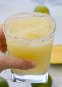 a hand holding a glass filled with an alcoholic drink next to some limes and bananas