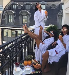 three women in white robes sitting on a balcony drinking orange juice and having breakfast together