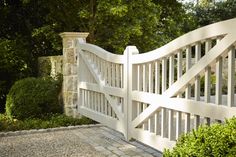 a white gate is in the middle of a stone walkway with bushes and trees behind it