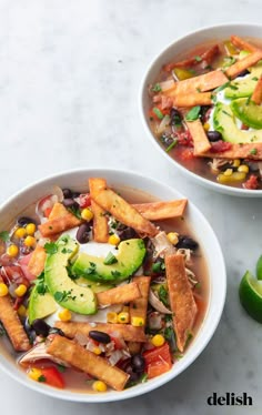 two white bowls filled with mexican soup and topped with tortilla chips, avocado, black beans, cilantro, lime wedges