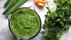 a blender filled with green liquid surrounded by fresh herbs and sliced bell peppers on a marble surface