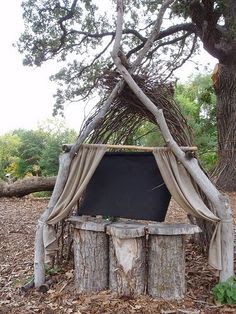 an outdoor shelter made out of tree trunks and branches, with a blackboard in the middle