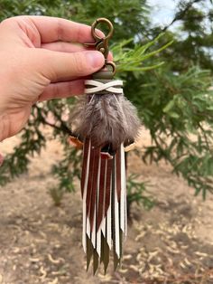 a hand holding a keychain with feathers attached to it's end, in front of a tree